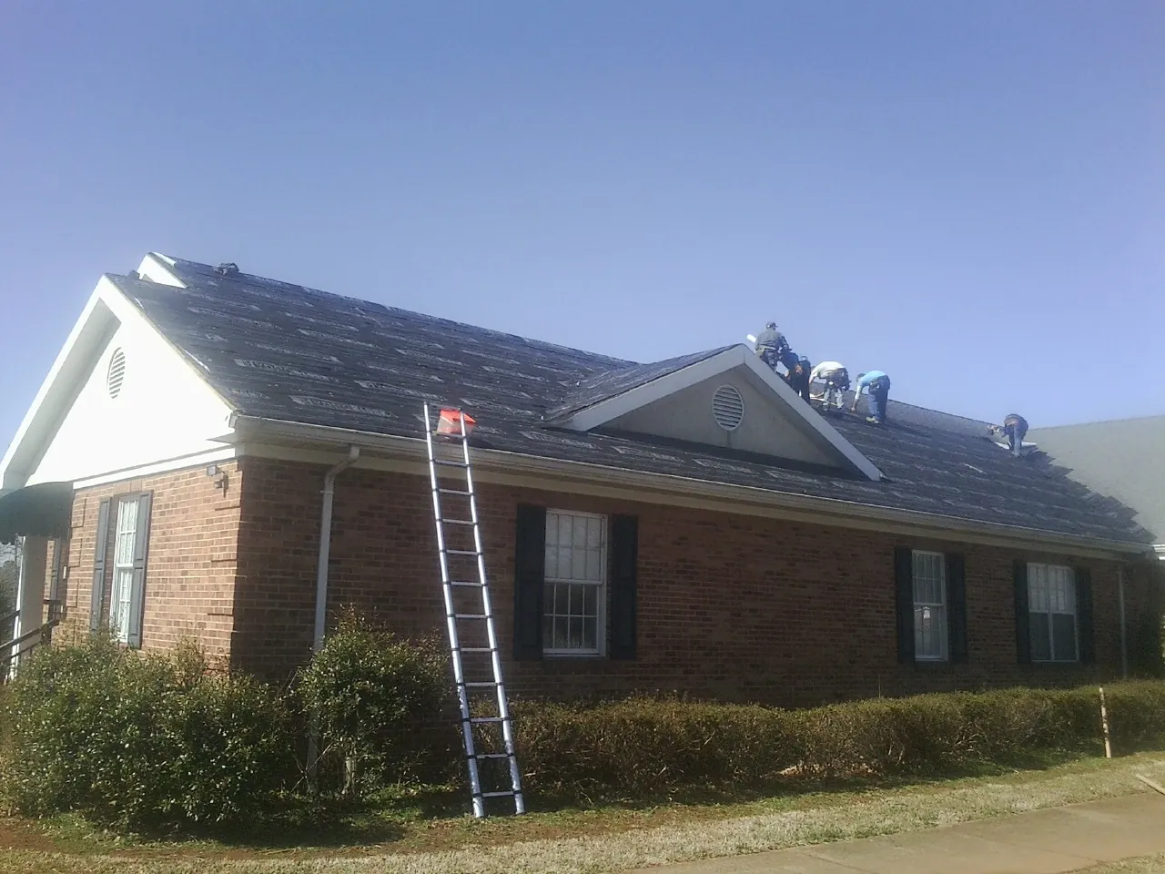 Crew tearing shingles off a brick ranch roof with a ladder set — Emergency Roof Repair work by our crew in Ivanhoe