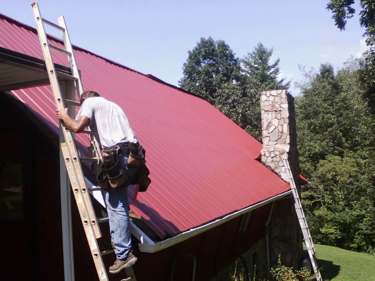 Roofer on a ladder beside a red metal roof and stone chimney — Soffit & Fascia Repair by Roofing Brothers in Ivanhoe